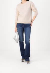 Beige gestrickter Pullover mit Raglanärmeln, kombiniert mit dunkelblauen Schlagjeans. Hält eine silberne metallische Clutch und trägt silberne Schuhe.