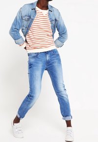 Chaqueta de mezclilla sobre un sudadera blanca y naranja a rayas, combinada con unos vaqueros claros desgastados y zapatillas blancas.