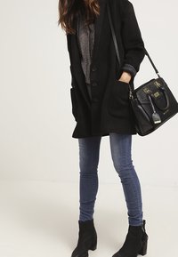 Kvinna klädd i svart kappa, mönstrad topp, blå skinny jeans, svarta ankelstövlar och bär en svart handväska med guldiga detaljer.