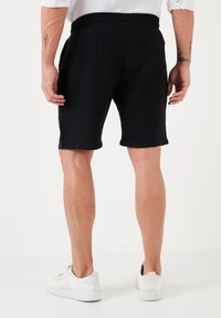 Zwarte gestructureerde shorts met een elastische tailleband, zijzakken en een achterzak, gecombineerd met witte sneakers op een effen achtergrond.