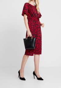 Punainen leopardi-printillä varustettu mekko, jossa on lyhyet hihat, kapea vyötärö ja polvenpituinen muotoilu. Yhdistetty mustan nahkalaukun ja mustien korkokenkien kanssa.