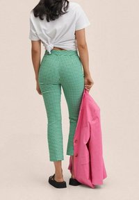 Pantalon à carreaux vichy verts avec une coupe ajustée, associé à un t-shirt blanc noué et un blazer rose, tenu d'une main. Chaussures plates noires.