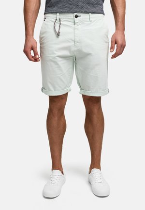 Mann trägt hellfarbene, hochgekrempelte Shorts mit seitlichen Taschen, Knopf- und Reißverschluss, kombiniert mit weißen Turnschuhen und einem grauen Hemd.