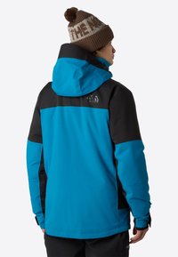 Blå och svart vattentät jacka med huva. Har en ficka med dragkedja och kontrasterande ärmpaneler. Bärs tillsammans med en brun stickad mössa.