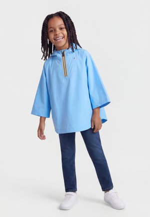 Hymyilevä lapsi, jolla on letitetyt hiukset, pukeutuneena vaaleansiniseen vetoketjulliseen ponchoon, tummiin farkkuihin ja valkoisiin tennareihin, seisoo yksinkertaisen taustan edessä.