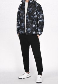 Veste noire pour homme avec motif abstrait bleu, fermeture éclair à l'avant, capuche, associée à un pantalon noir et des baskets blanches. Design moderne avec un look décontracté.