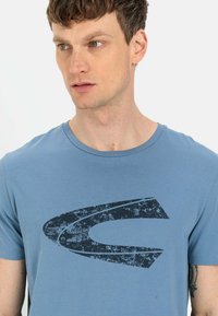 Himmelblaues T-Shirt mit kurzen Ärmeln aus weichem Stoff, das auf der Vorderseite ein großes, abgenutztes schwarzes Grafikdesign zeigt.