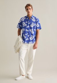 Camisa azul de manga corta con un patrón floral blanco, combinada con pantalones de color claro y zapatillas blancas.