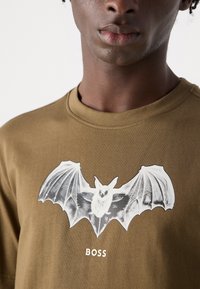 Olive green t-shirt made of cotton featuring a white bat graphic and "BOSS" text below. Simple crew neck design and soft texture.