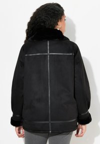 Giacca nera in microfibra scamosciata con colletto in pelliccia sintetica, vestibilità oversize e dettagli in pelle sulle cuciture. Presenta una superficie liscia e maniche lunghe.