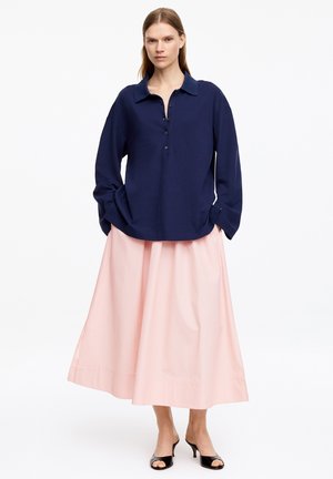Žena stojaca, oblečená v námornícky modrej košeli na gombíky s dlhými rukávmi, svetloružovej midi sukni a čiernych topánkach na podpätku s otvorenou špičkou, na bielom pozadí.