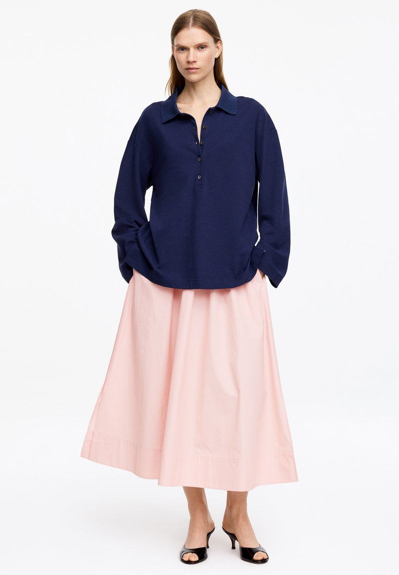 Femme debout portant une chemise bleu marine à manches longues boutonnée, une jupe midi rose clair et des talons ouverts noirs sur fond blanc.