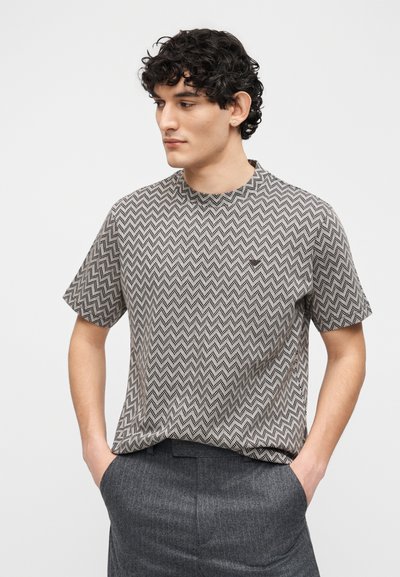 Νεαρός με σγουρά σκούρα μαλλιά, ντυμένος με πουκάμισο κοντομάνικο με ζικ-ζακ μοτίβο και σκούρο γκρι παντελόνι, με τα χέρια στις τσέπες, κοιτάζει προς τα αριστερά.