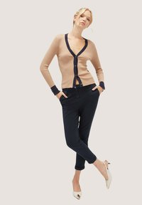 Beige žebrovaný cardigan s námořnickým lemováním a knoflíky, spárovaný s tmavými kalhotami. Modelka v bílé slingback obuvi, pózující s rukama v kapsách.