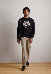 Schwarzer Sweatshirt mit Tiger-Graphic, beiger Hose und schwarzen Schuhen. Model mit kurzem Haar steht auf einem Holzfußboden vor einem neutralen Hintergrund.