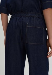 Dark blue denim pants featuring an elastic waistband, a single back pocket with beige stitching, and a loose-fitting design.