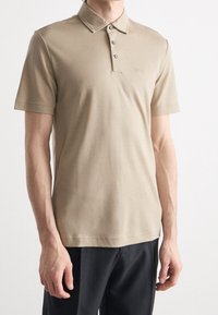 Beige polo shirt featuring a classic collar, three-button placket, short sleeves, and subtle branding embroidered on the chest.