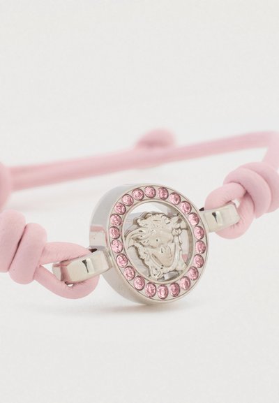 Rožinės odos apyrankė su centre esančia metaline medaliuote, išgraviruotais raštais ir apsupta rožinių dekoratyvinių akmenų.