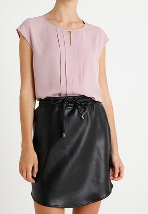 Femme portant un chemisier sans manches rose clair avec des plis et une jupe en cuir noire avec une taille à cordon.
