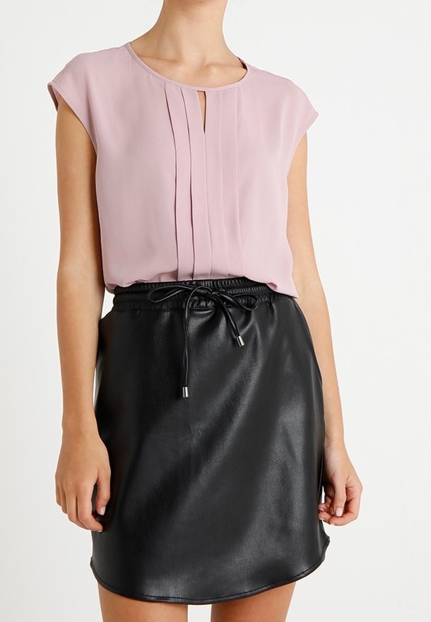 Femme portant un chemisier sans manches rose clair avec des plis et une jupe en cuir noire avec une taille à cordon.