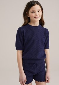 Tumes sinine koonuseline lühikeste varrukatega topp, millel on tekstuuriga diamantruut ja soonikkoes krae, koos sobivate tekstuuriga lühikeste pükstega, millel on laineline äär.