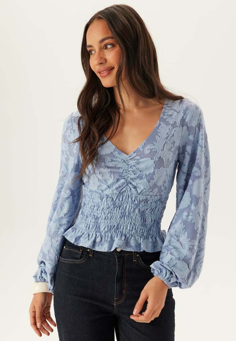 Kvinde med langt mørkt hår iført en lyseblå, blomstertekstureret bluse med lange ærmer og rynket talje samt mørke jeans, vendt mod venstre og smilende.