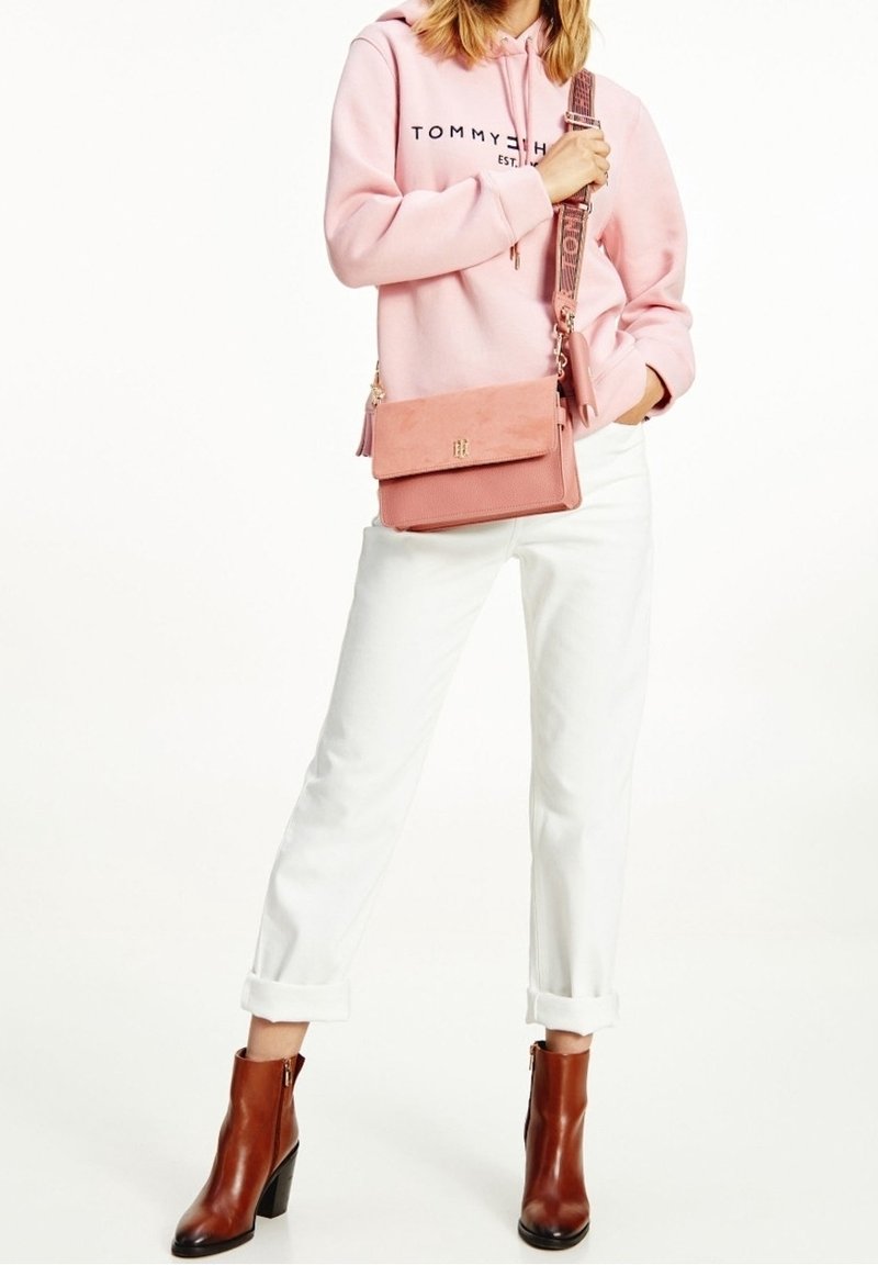 Růžová semišová crossbody kabelka s texturovaným povrchem, zlatými kovovými detaily a vzorovaným popruhem, v kombinaci se světle růžovou mikinou a bílými džíny.