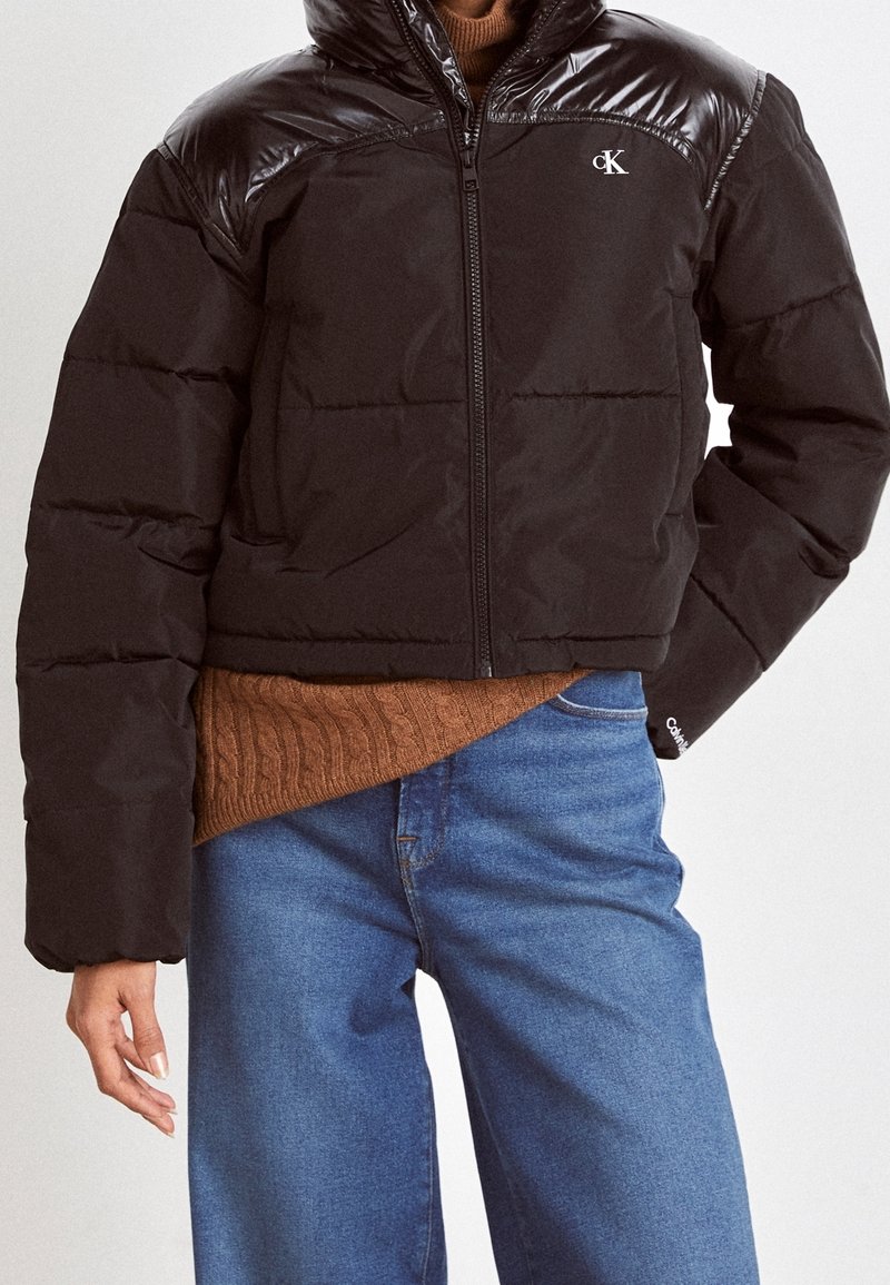 Giacca puffer nera corta con dettagli lucidi sulle spalle, zip completa e logo Calvin Klein. Abbinata a un maglione lavorato marrone e jeans blu.