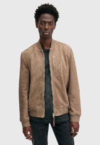 Giacca bomber in suede color tan con zip frontale, colletto e polsini a coste, tasche laterali e vestibilità slim. Indossata sopra una maglietta nera e jeans.