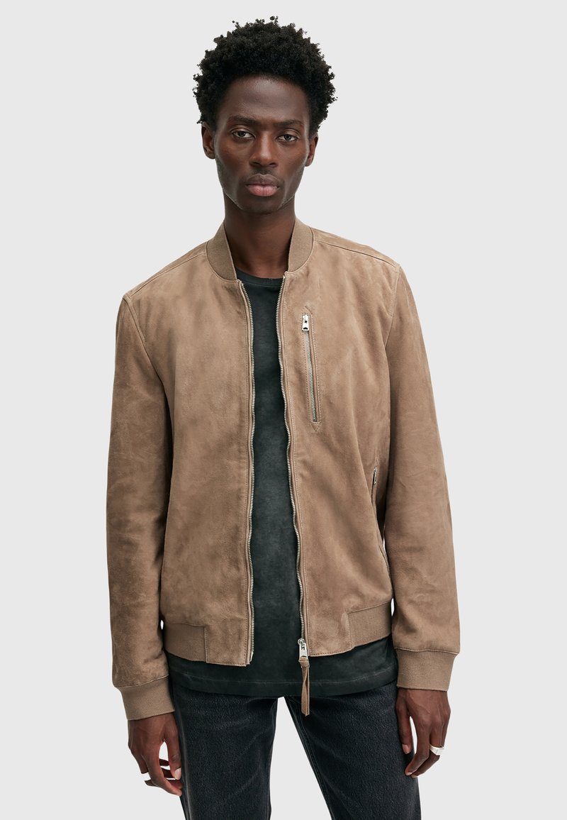 Giacca bomber in suede color tan con zip frontale, colletto e polsini a coste, tasche laterali e vestibilità slim. Indossata sopra una maglietta nera e jeans.
