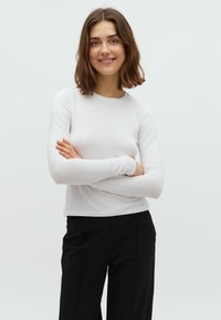 Balta garām piedurknēm krekls ar apaļu izgriezumu. Mīksts, piegulošs audums, minimālistisks dizains, apvienots ar melnām biksēm. Modelis pozē ar krustotiem rokām.