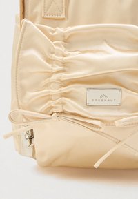 Μπεζ σακίδιο πλάτης με λαμπερή σατέν υφή, μεταλλική πλακέτα λογότυπου με την ένδειξη "DOUGHNUT" και κορδόνια για ρύθμιση στη βάση.