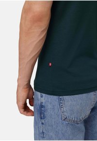 Dunkelgrünes Baumwoll-T-Shirt mit kurzen Ärmeln, das am Saum ein kleines rotes Flaggenakzent hat. Getragen mit hellblauen Jeans.