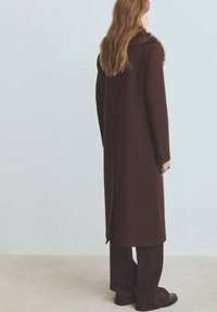 Bruine wollen jas met een nepbonten kraag en een lange, rechte silhouet. De jas wordt aangevuld met bijpassende bruine broek met wijde pijpen.