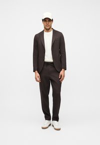 Homme portant un blazer marron foncé et un pantalon assorti, avec un sweat-shirt blanc, une casquette blanche et des baskets blanches, debout devant un fond uni.