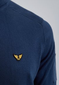 Dunkelblau gestrickter Pullover mit einem runden Halsausschnitt und einem kleinen goldenen geflügelten Emblem auf der linken Brust. Weiche Textur mit Rippdetails.