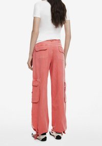 Růžové cargo kalhoty vyrobené z lehkého materiálu, s velkými bočními kapsami, nastavitelné nohavice a volný střih. Zobrazeno ze zadní strany s bílým topem.