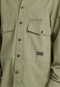 Oliwkowa koszula zapinana na guziki z dwiema przednimi kieszeniami. Posiada brązowe guziki oraz metkę Timberland na dolnej kieszeni. Miękka faktura materiału.