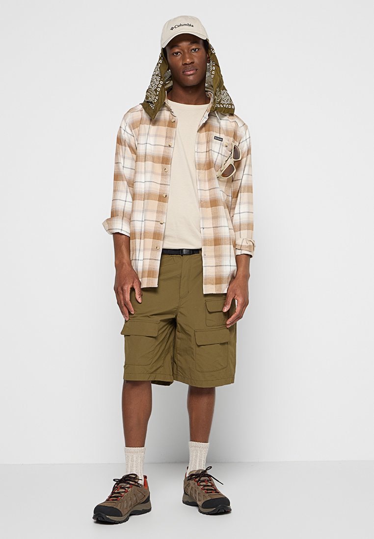 Joven con gorra beige y bandana, camisa de cuadros en tonos marrones sobre camiseta beige, pantalones cortos de carga verde oliva, zapatos de senderismo y calcetines, de pie contra una pared blanca.