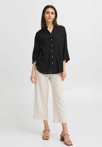 Maglietta nera con bottoni, dalla vestibilità comoda, maniche arrotolate e bottoni in ottone. Abbinata a pantaloni beige chiaro a gamba larga e sandali con tacco marroni.