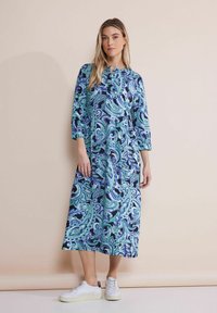 Midi šaty s paisley vzorem v odstínech modré a zelené, s rukávy ke třetině, kulatým výstřihem; model má na sobě bílé tenisky.