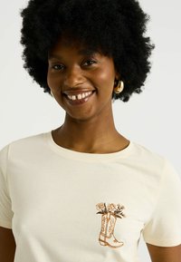 Camiseta de algodón crema con un diseño bordado marrón de una bota de cowboy con flores. Cuello redondo y mangas cortas.