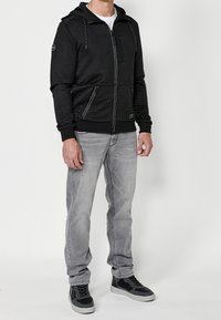 Giacca nera con cappuccio e zip, con texture a coste, tasche frontali con zip e coulisse bianche a contrasto. Indossata sopra jeans grigio chiaro e sneaker nere.