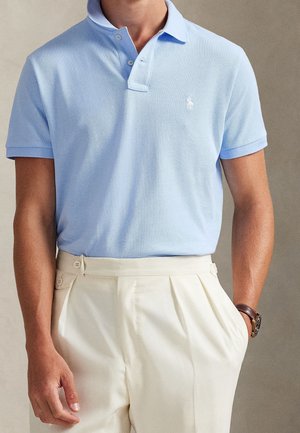 Homme portant un polo bleu clair à manches courtes rentré dans un pantalon plissé crème taille haute avec une montre-bracelet marron.