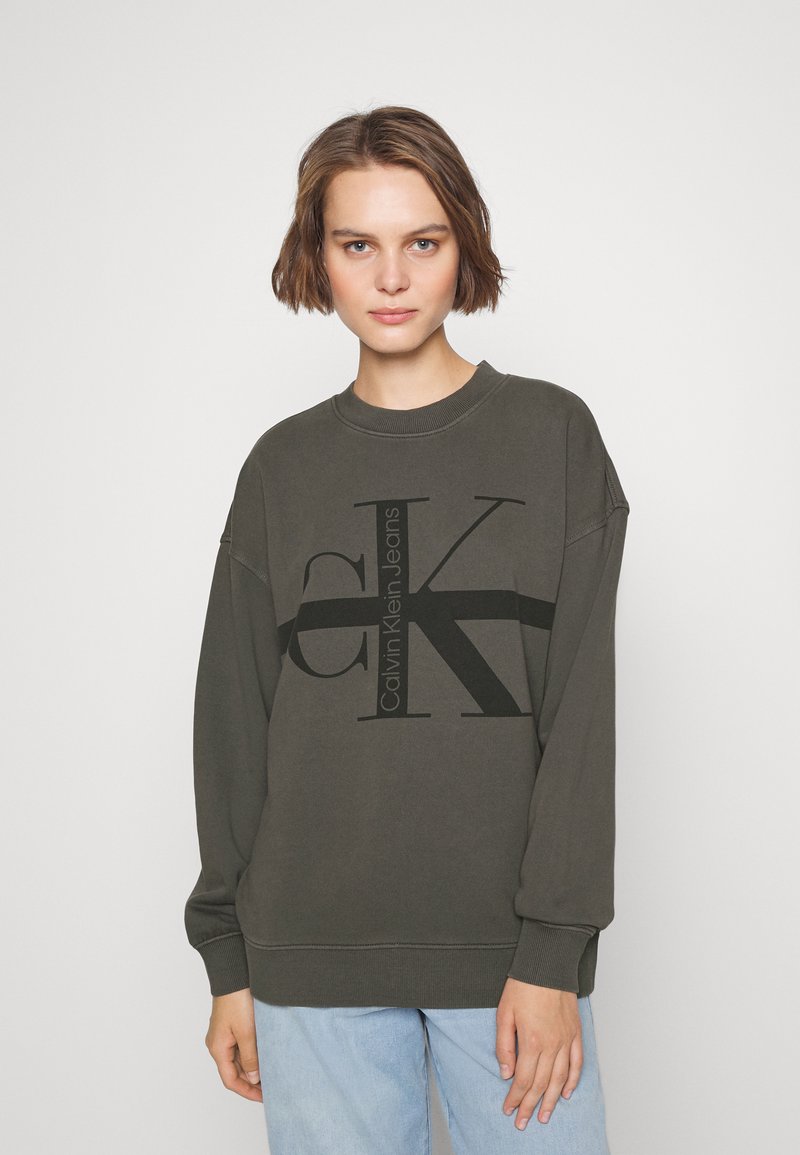 Sudadera verde oscura con cuello redondo, de corte oversized, que presenta un gran logo gráfico en negro 'CK' y el nombre de la marca en el frente.