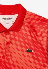 Červené polo tričko s texturovaným geometrickým vzorem, klasickým límečkem a třemi knoflíky. Logo Lacoste je umístěno na levé straně.