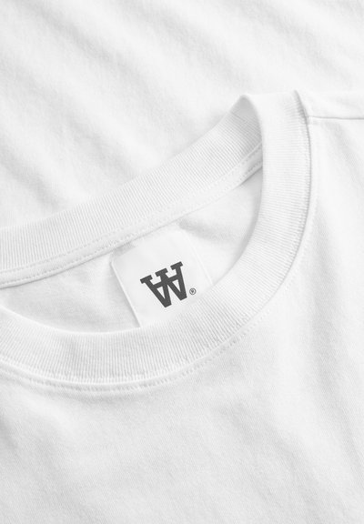 T-shirt bianco in cotone con scollo rotondo a coste, dotato di un'etichetta con un logo scuro. La texture appare liscia e morbida.