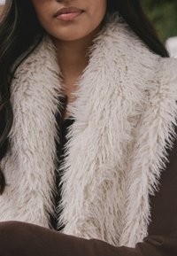  Kunstpelzschal in Creme mit einer zotteligen Struktur, geschichtet über einem schokoladenbraunen Oberteil. Der Schal zeichnet sich durch ein weiches, voluminöses Design aus.