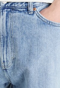Acercamiento de un bolsillo de unos jeans de mezclilla azul claro con la mano parcialmente dentro, mostrando el detalle de la costura y el remache de cobre.