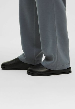 Black footwear with a smooth finish, rounded toes, and minimalistic design. Paired with loose-fitting, light gray pants with stitched accents.